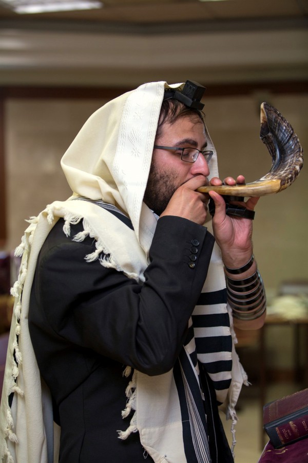 A Jewish man wearing a tallit (prayer shawl) and tefillin (phylacteries) blows a shofar (ram's horn).